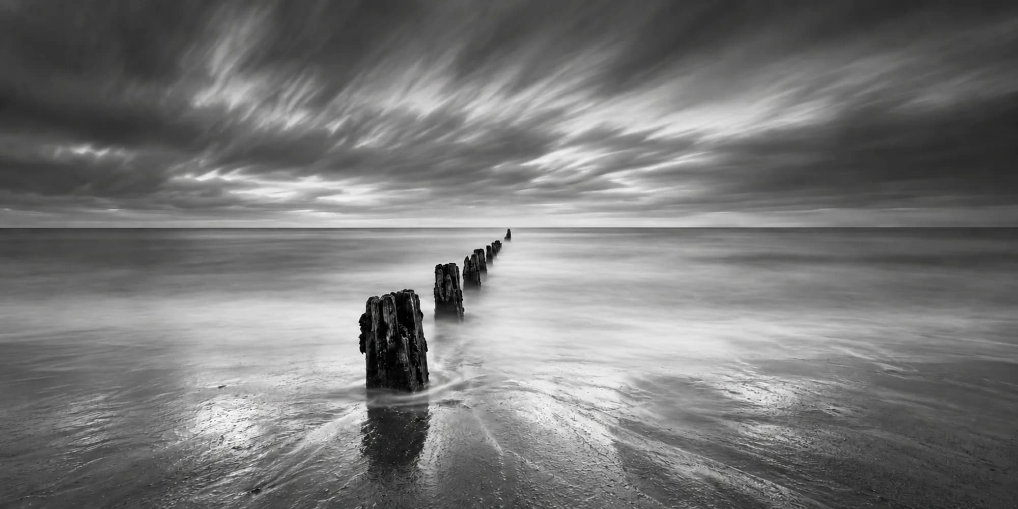 Black-and-white long exposure of a line of wooden posts leading into a smooth sea