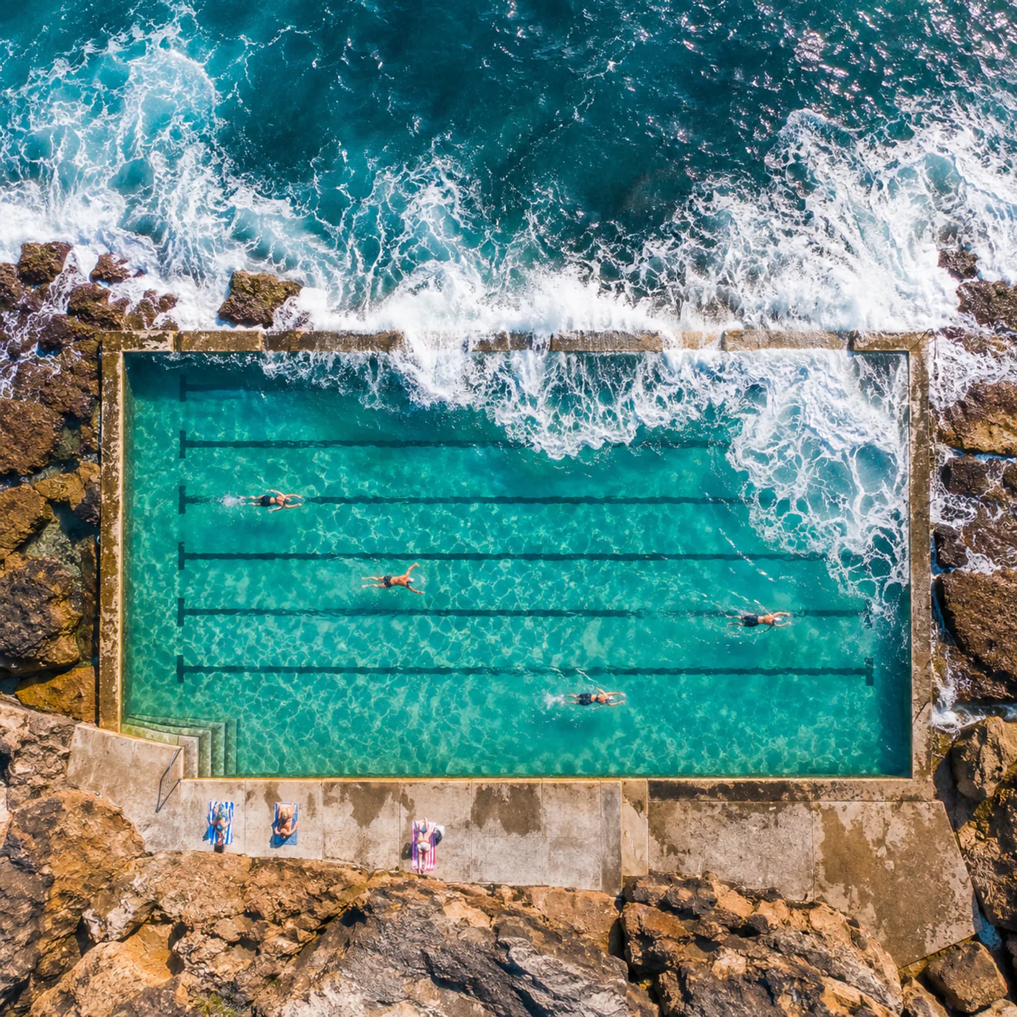 A rectangular ocean pool cut into rocks with waves breaking over its edge, seen from above